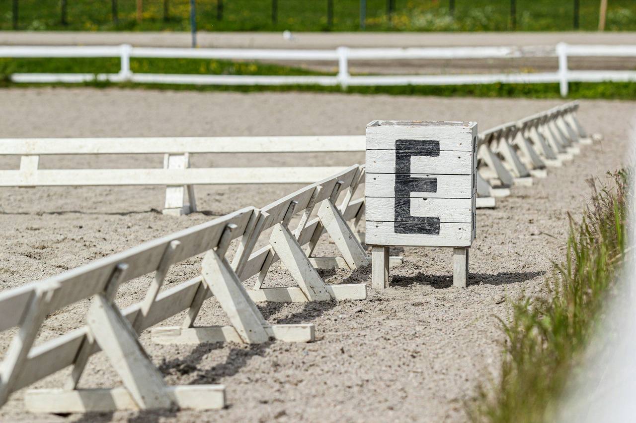 An empty sand dressage arena edged with white boards, with the letter E marker in the foreground