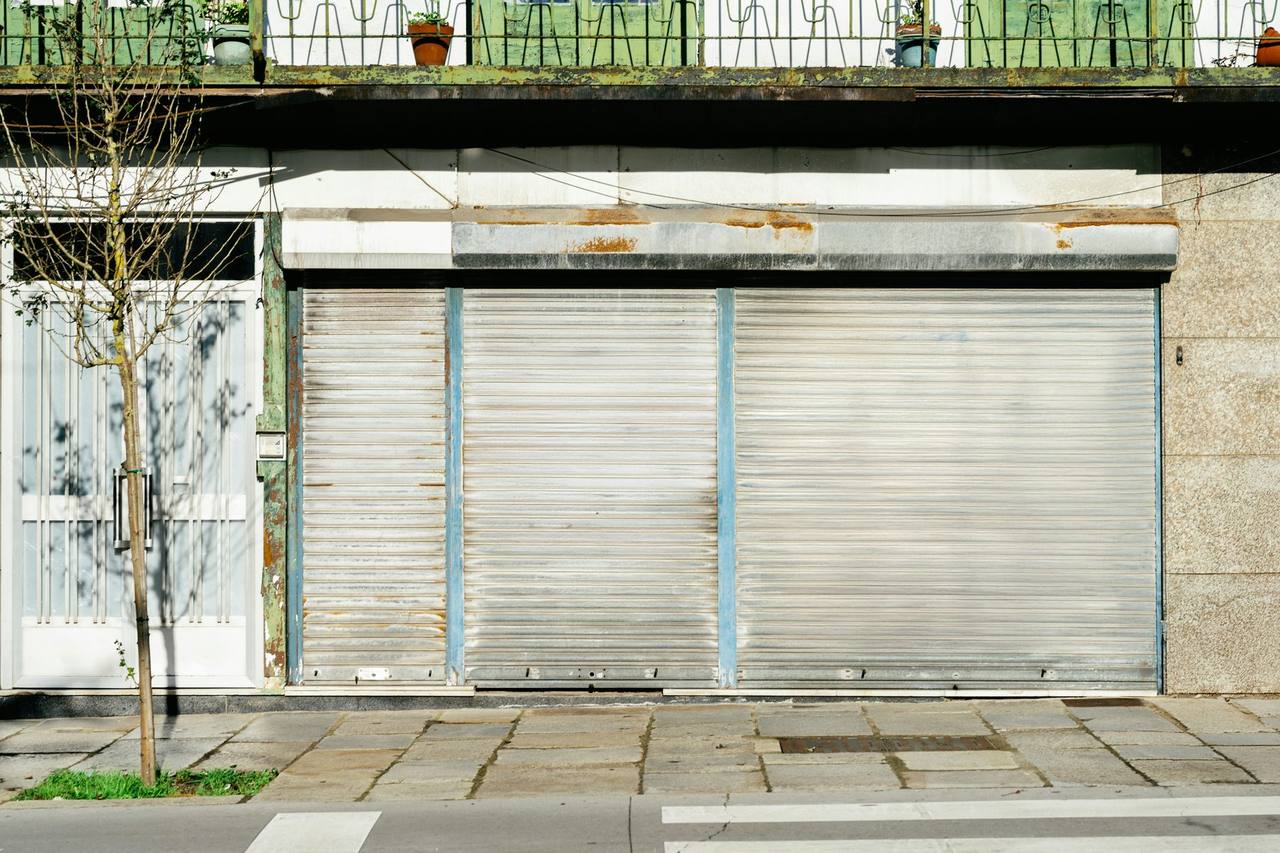 A closed ground floor shop unit with grey roller shutters pulled down on a quiet street