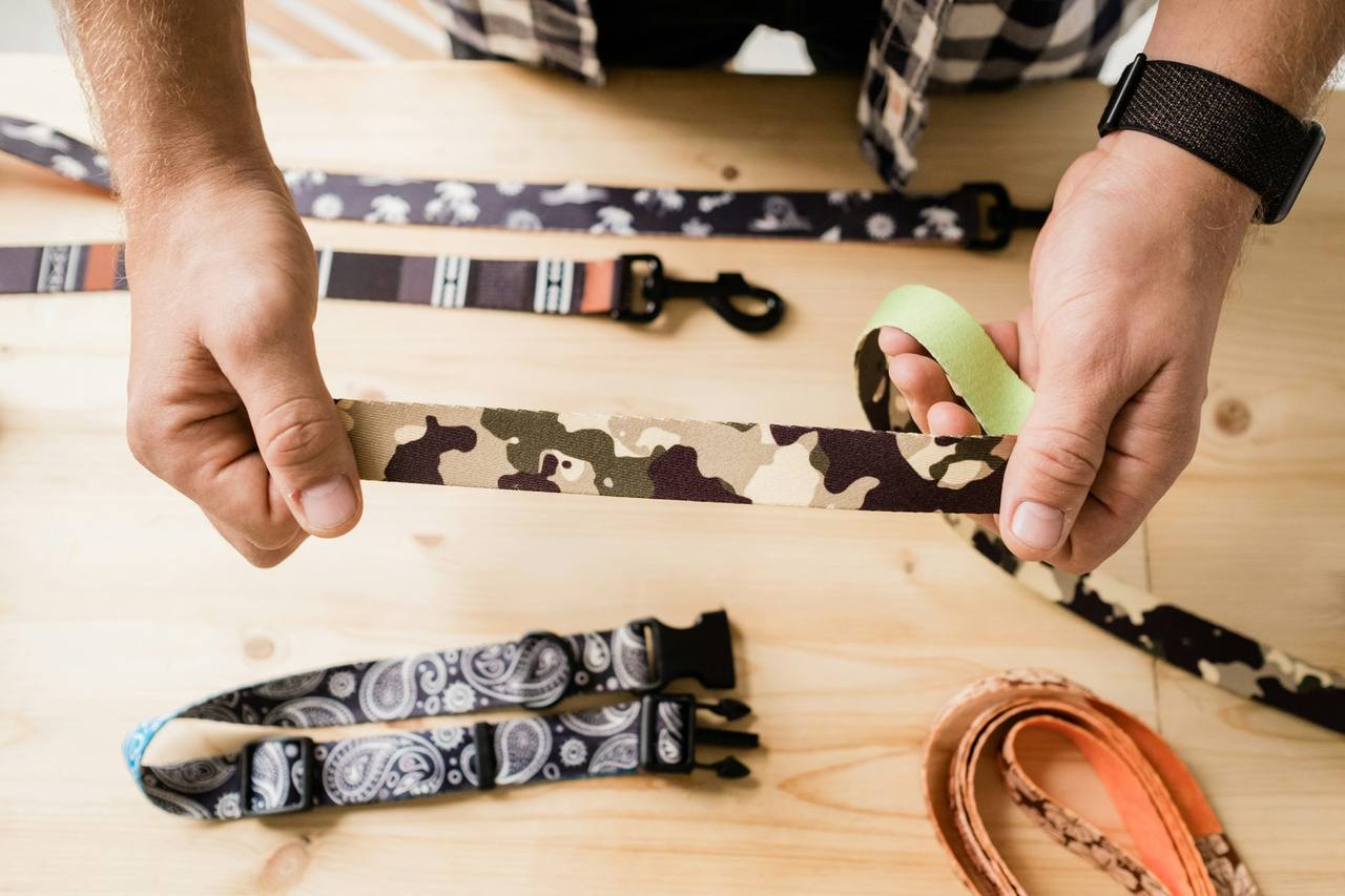 Hands holding a camouflage print dog lead above patterned collars and leads laid on a pine table