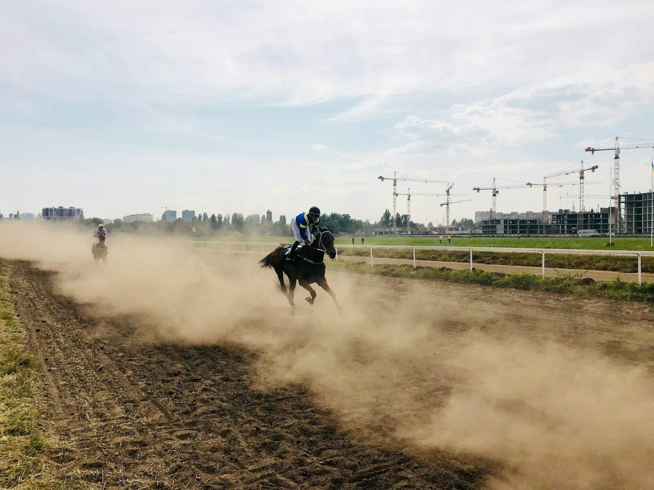 A jockey on a dark racehorse galloping on a dusty dirt track, with construction cranes in the distance