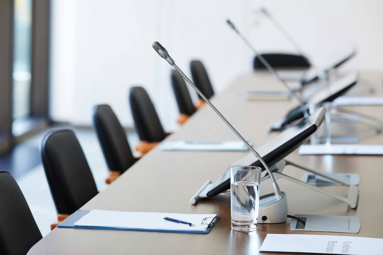 An empty boardroom table with black chairs, desk microphones, papers and glasses of water
